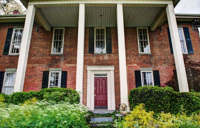 A red-brick house with a maroon door, white columns, green shrubs, and yellow flowers in front—this charming property offers inviting bed and breakfast rooms in Springfield, Ohio. Two lion statues sit on either side of the steps leading to the entrance.