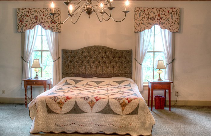 A neatly made bed with a patterned quilt is centered between two windows, each with a side table and lamp—an inviting scene reminiscent of charming bed and breakfast rooms in Springfield, Ohio. A chandelier hangs above, and curtains with valances frame the windows.