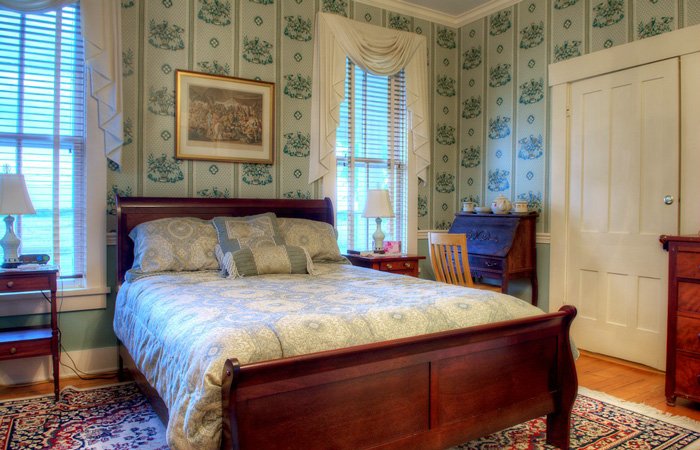 A cozy bedroom reminiscent of bed and breakfast rooms in Springfield, Ohio, featuring a wooden bed, patterned bedding, vintage wallpaper, two windows, a desk with a chair, bedside tables with lamps, a framed picture, and a patterned rug.