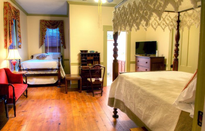 A cozy bedroom with a canopy bed, a single bed, wooden floor, desk, TV, red loveseat, and a lamp by the window with patterned curtains—perfect for guests searching for charming bed and breakfast rooms in Springfield, Ohio.
