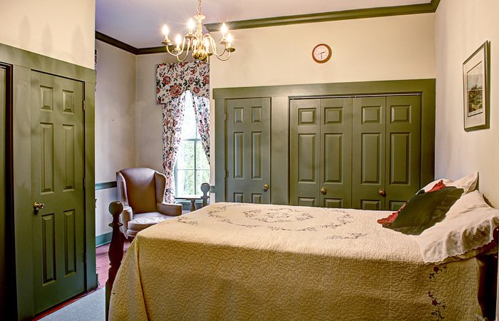 A cozy bedroom with green-trimmed double doors, a vintage chandelier, floral curtains, an armchair by the window, and a bed with a cream quilt and accent pillows—just one of the inviting bed and breakfast rooms in Springfield, Ohio. A clock and framed art hang on the cream walls, creating a charming retreat.