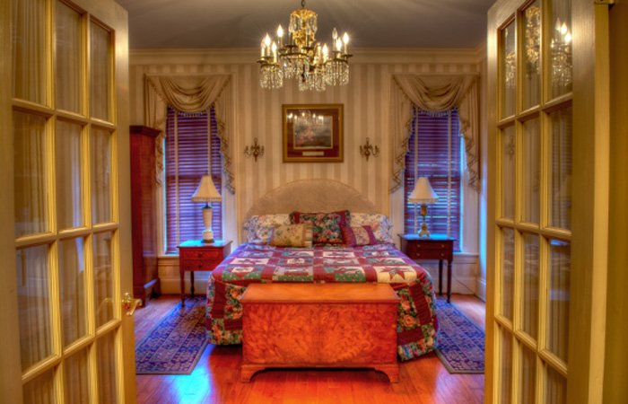 A warmly lit bedroom with a large bed covered in a patchwork quilt, two side tables with lamps, a chandelier, and two windows with drapes. Reminiscent of charming bed and breakfast rooms in Springfield Ohio, the inviting space is viewed through open French doors.