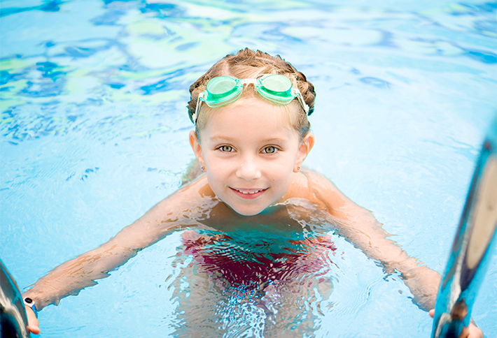 A young girl with green goggles on her head smiles while holding onto the edge of a swimming pool, partially submerged in clear blue water—a perfect example of the fun things to do in Springfield, Ohio.