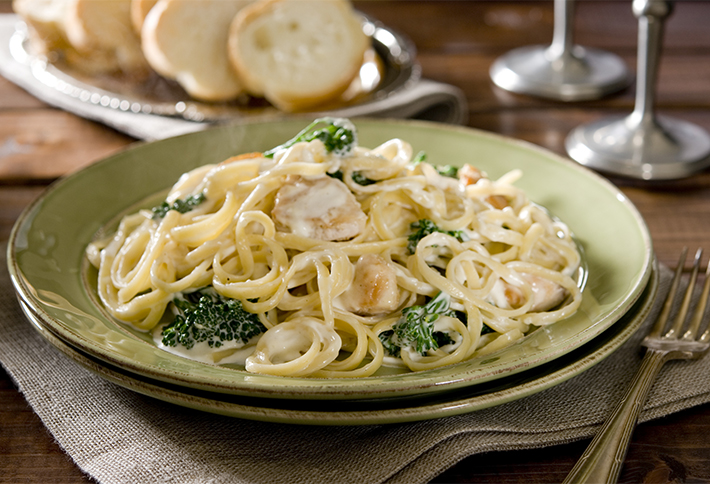 A plate of creamy pasta with chicken and broccoli sits on a green dish, with slices of bread and two wine glasses in the background. A fork and knife rest beside the plate on a cloth napkin, making it a perfect meal to enjoy after exploring things to do in Springfield, Ohio.