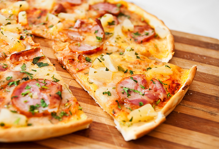 Thin crust pizza topped with cheese, ham, pineapple chunks, herbs, and tomato sauce, sliced on a wooden cutting board—a perfect treat to enjoy after exploring the top things to do in Springfield, Ohio.
