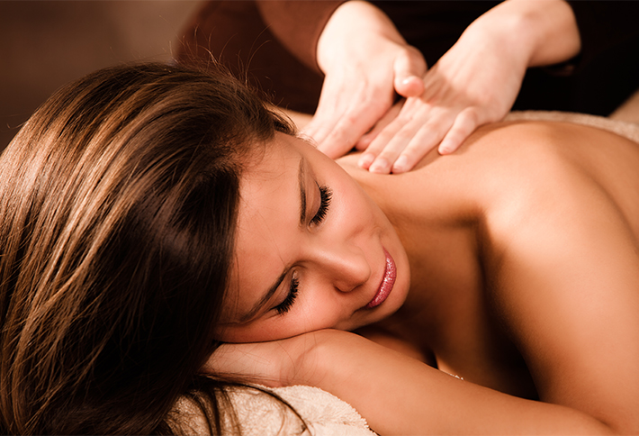 A woman with long brown hair lies on her stomach with eyes closed, resting her head on a towel while receiving a back and shoulder massage from another person. The setting appears warm and relaxing, making it an ideal choice among the many things to do in Springfield, Ohio for those seeking tranquility and rejuvenation.