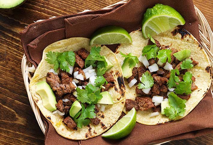 Two beef tacos topped with avocado, onions, and cilantro sit on a brown napkin in a basket. Lime wedges are placed beside and on the tacos—making this fresh dish a delicious highlight among the many things to do in Springfield, Ohio.