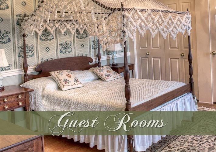 Elegant guest bedroom with a wooden four-poster bed, lace canopy, floral wallpaper, and decorative pillows. Overlay text reads “Guest Rooms.”.